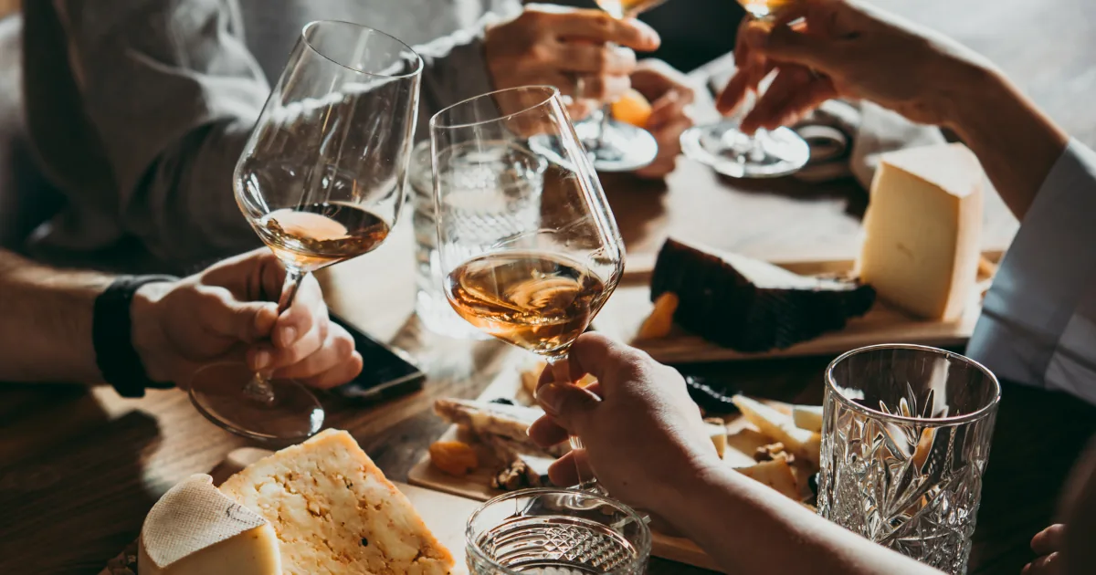 Hands over a cheese board, holding glasses of amber wine in daylight.